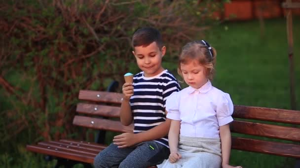 Niño comiendo un helado y sentado en el banco mientras que la chica está buscando — Vídeo de stock