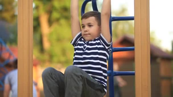 Um garotinho em uma camiseta listrada está brincando no playground, balançando em um balanço.Primavera, tempo ensolarado — Vídeo de Stock