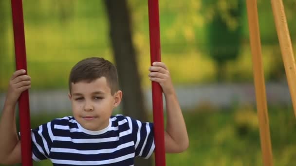 En liten pojke i en randig T-shirt spelar på lekplatsen, gunga på en gunga. Våren, soligt väder — Stockvideo