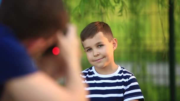 Маленький хлопчик в смугастій футболці посміхається і веселить, поки фотограф робить фото. Весна, сонячна погода — стокове відео