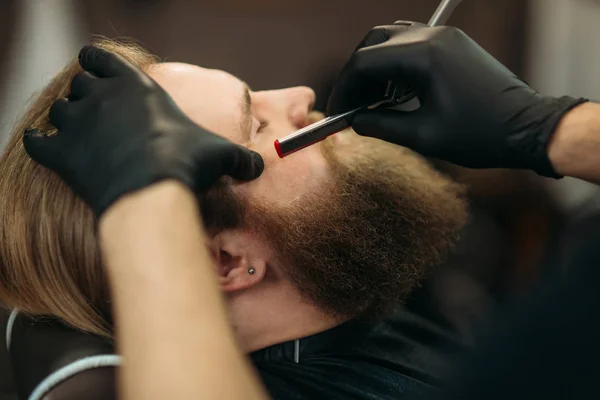 Bebaarde man met lange baard om stijlvolle haar scheren, kapsel, met scheermes van barber in de barbershop — Stockfoto