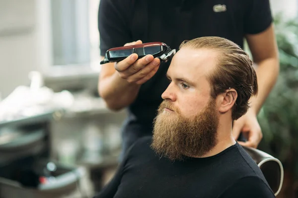Jóképű férfi fodrász levágatja a haját, miközben székben ül a fodrászatban. — Stock Fotó