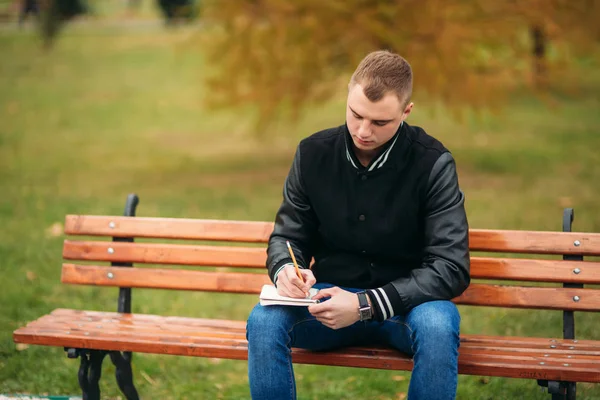 穿着黑色夹克的学生坐在长凳上的公园里, 把他的想法写在笔记本上。英俊的男孩 — 图库照片