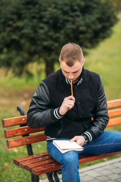 Student v černá bunda sedí v parku na lavičce napíše své myšlenky v poznámkovém bloku. Hezký kluk — Stock fotografie