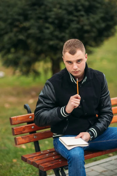 Student v černá bunda sedí v parku na lavičce napíše své myšlenky v poznámkovém bloku. Hezký kluk — Stock fotografie