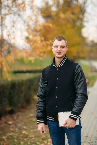 Stylish guy in black jucket stand in park. He hold a notebook in hands — Stock Photo, Image