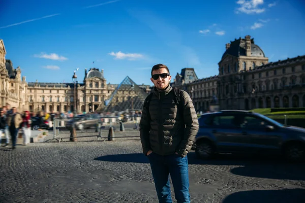 Un joven turista con chaqueta caqui está de pie en el fondo del Louvre. Tiempo soleado de otoño — Foto de Stock
