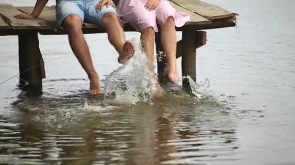 Beautiful youn couple sit on the pier near the lake. make splash by feet. slow motion — Stock Video