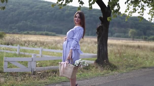 Schöne schwangere Frau in blauem Kleid, die den Hof betritt. Stricktasche mit Blumenstrauß. Hintergrund von Bäumen und Bergen. Zeitlupe — Stockvideo