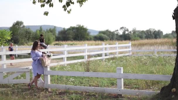 Mulher grávida bonita em vestido azul andando ouvir a fazenda. Saco de malha com buquê de flores. Antecedentes de árvores e montanhas. Movimento lento — Vídeo de Stock