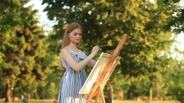 Menina bonita de pé no parque e desenha um quadro usando uma paleta com tintas e uma espátula . — Vídeo de Stock