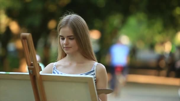 Menina bonita de pé no parque e desenha um quadro usando uma paleta com tintas e uma espátula . — Vídeo de Stock