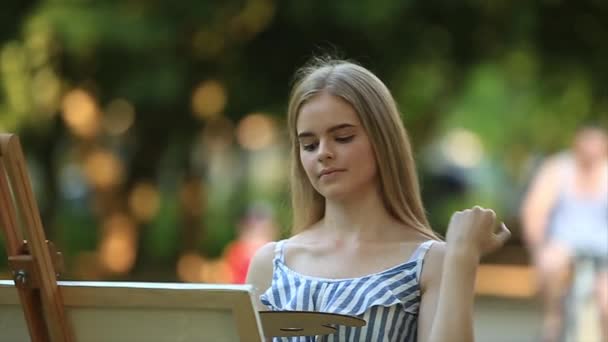 Menina bonita de pé no parque e desenha um quadro usando uma paleta com tintas e uma espátula . — Vídeo de Stock