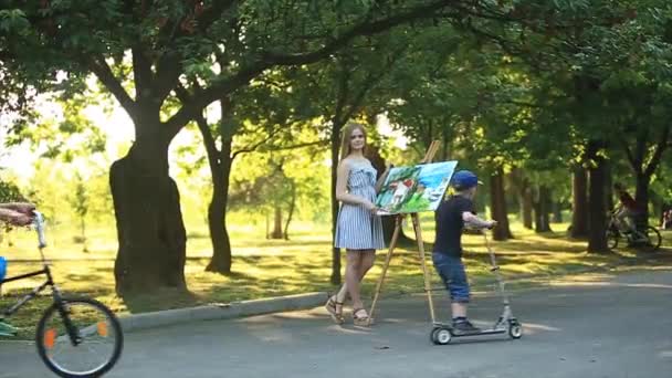 Hermosa chica de pie en el parque y dibuja un cuadro usando una paleta con pinturas y una espátula . — Vídeos de Stock