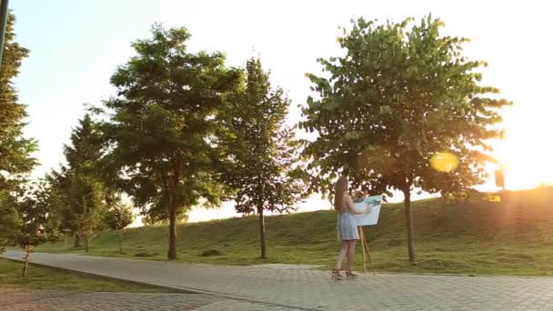Menina bonita desenha um quadro no parque usando uma paleta com tintas e uma espátula. Easel e lona com uma foto. O verão é um dia ensolarado, pôr do sol . — Vídeo de Stock