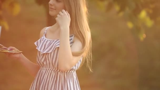 Vacker flicka ritar en bild i parken med hjälp av en palett med färger och en spatel. Staffli och duk med en bild. Sommaren är en solig dag, solnedgång. — Stockvideo