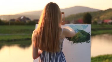 Güzel kız parkta boyalar ve bir spatula ile bir palet kullanarak bir resim çizer. Şövale ve tuval resmi. Güneşli bir gün, gün batımı yaz mi.