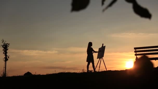 La silhouette di una ragazza bionda dipinge un dipinto sulla tela con l'aiuto di vernici. Un cavalletto di legno mantiene il quadro. L'estate è una giornata di sole, tramonto — Video Stock