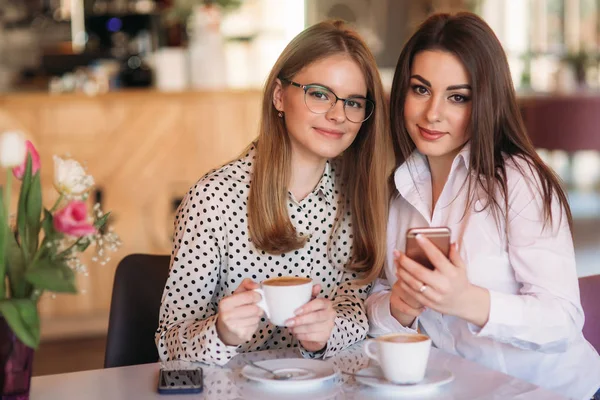 Dziewczyny, siedząc w kawiarni picia cappuccino i za pomocą telefonów — Zdjęcie stockowe