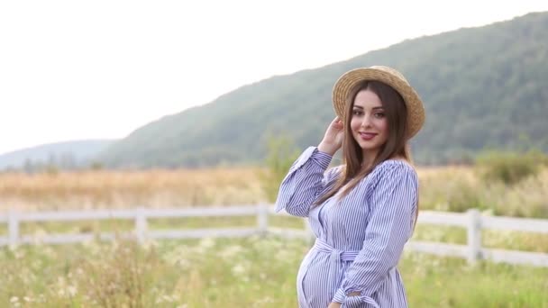 La mujer embarazada en la naturaleza se prueban el sombrero de punto. Antecedentes de campo y fachada blanca cerca de la granja. montañas — Vídeo de stock