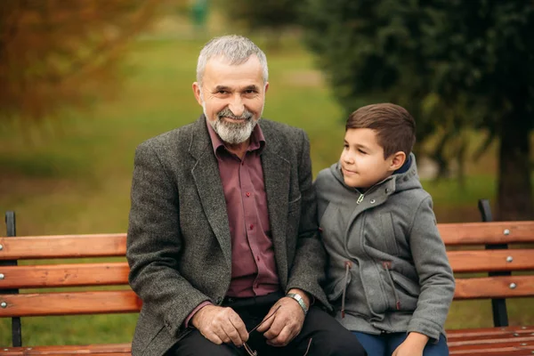 爷爷和他的孙子一起在公园里消磨时间。他们坐在长椅上互相看着对方 — 图库照片
