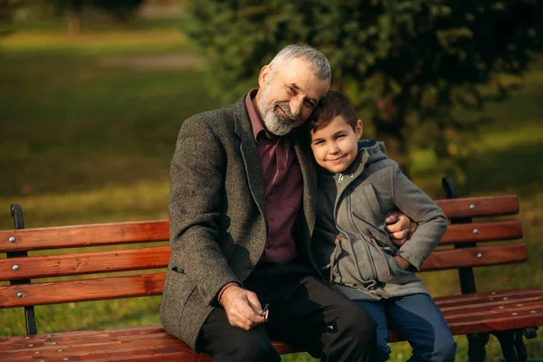 爷爷和他的孙子一起在公园里消磨时间。他们坐在长椅上互相拥抱 — 图库照片