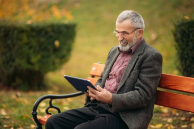 Büyükbaba bir tablet kullanır ve parkta bankta oturur. O ekrana bak