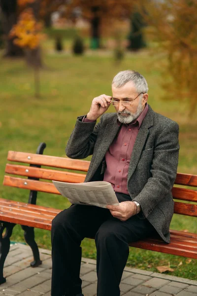 Avô bonito com uma bela barba em uma jaqueta cinza senta-se em um banco no parque e lê um jornal — Fotografia de Stock