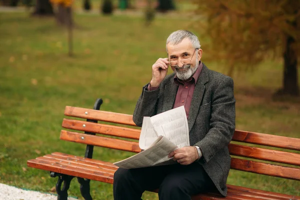 Stilig farfar med vackra skägg i en grå jacka sitter på en bänk i parken och läser en tidning — Stockfoto