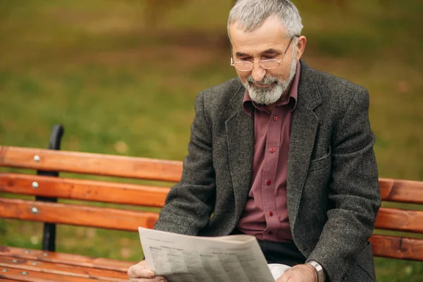 Красивый дедушка с красивой бородой в серой куртке сидит на скамейке в парке и читает газету — стоковое фото