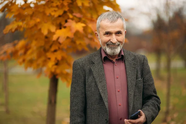 Un bell'uomo anziano con la barba negli occhiali sta usando un telefono. Passeggiata nel parco in autunno — Foto Stock