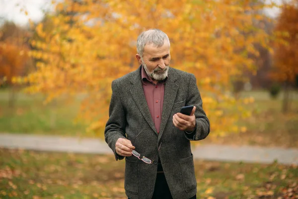 Ένας όμορφος ηλικιωμένος άνδρας με γενειάδα σε ποτήρια χρησιμοποιώντας ένα τηλέφωνο. Βόλτα στο πάρκο το φθινόπωρο — Φωτογραφία Αρχείου