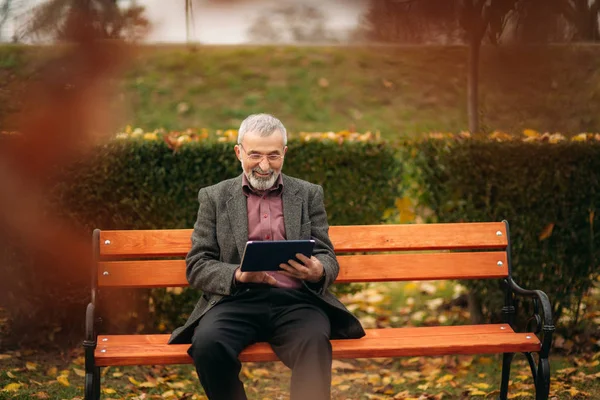 O avô usa um tablet e senta-se no parque no banco. Ele olha para o ecrã — Fotografia de Stock