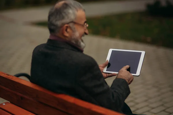 El abuelo usa una tableta y se sienta en el parque en el banco. Él mira a la pantalla y mira la foto —  Fotos de Stock