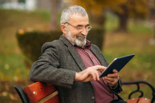 El abuelo usa una tableta y se sienta en el parque en el banco. Él mira a la pantalla y mira la foto —  Fotos de Stock