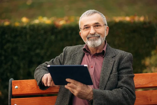 Nagypapa használ egy tablettát, és ül a parkban a padon. Úgy néz ki, hogy a kamera és a mosoly — Stock Fotó
