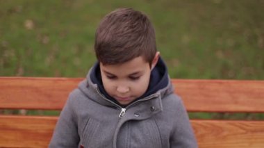Küçük çocuk parkı ve gülümseme kamera için bankta oturmuş. Yakışıklı çocuk ceketli. sonbahar. ağır çekim