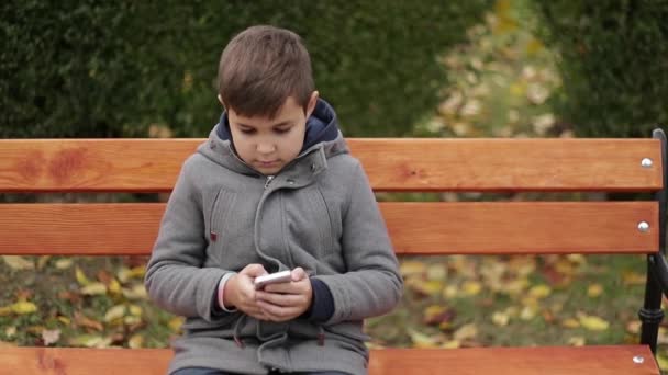 Kleiner Junge benutzt ein Handy im Park. Bei Herbstwetter sitzt er auf der Bank. gelbe Bäume auf dem Hintergrund — Stockvideo