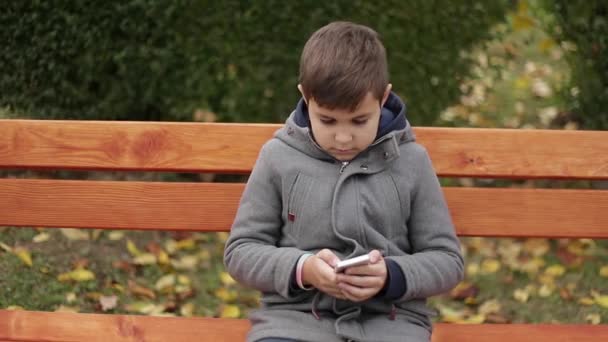 Kleiner Junge benutzt ein Handy im Park. Bei Herbstwetter sitzt er auf der Bank. gelbe Bäume auf dem Hintergrund — Stockvideo