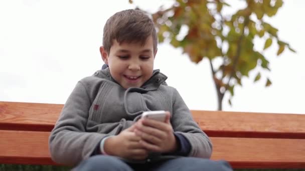 Kleine jongen een telefoon gebruiken in het park. Hij zit op de Bank in herfstweer. Gele bomen op de achtergrond — Stockvideo
