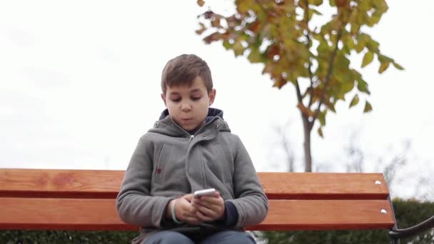 El niño usa un teléfono en el parque. Se sienta en el banco en otoño. Árboles amarillos sobre fondo — Vídeos de Stock