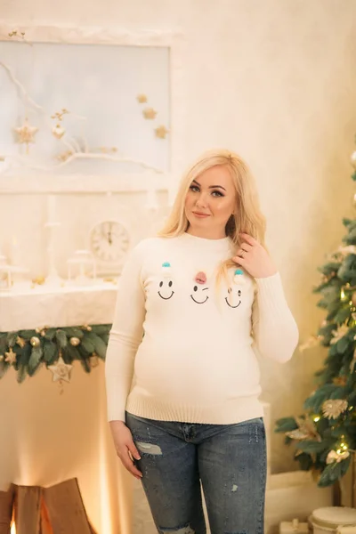 Красивая беременная блондинка в рождественском свитере стоит возле рождественской елки — стоковое фото