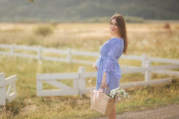 Mulher grávida bonita em vestido azul andando ouvir a fazenda. Saco de malha com buquê de flores. Fundo de árvore e montanhas — Fotografia de Stock