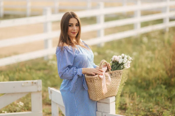 Schöne schwangere Frau in blauem Kleid, die den Hof betritt. Stricktasche mit Blumenstrauß. Hintergrund von Baum und Bergen — Stockfoto