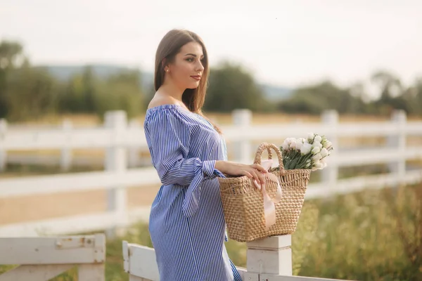 Belle femme enceinte en robe bleue marchant entendre la ferme. Sac tricoté avec bouquet de fleurs. Contexte de l'arbre et des montagnes — Photo