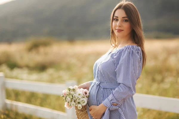 Vista de cerca de la hermosa mujer embarazada en el campo sosteniendo ramo y sonrisa. Relájate en la naturaleza. Ella puso su mano sobre su vientre —  Fotos de Stock