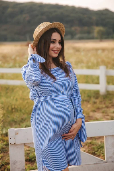 Femme enceinte dans la nature essayer chapeau tricoté. Fond de champ et clôture blanche près de la ferme — Photo