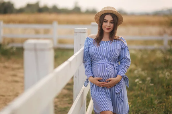 Hermosa mujer embarazada en vestido azul y sombrero de punto de pie cerca de la novia en la granja. Ella puso las manos sobre su vientre y sonrió —  Fotos de Stock