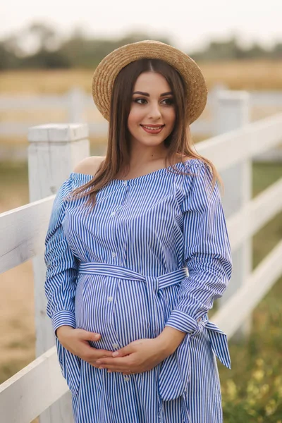 Belle femme enceinte en robe bleue et chapeau tricoté debout près de la clôture dans la ferme. Elle mit les mains sur son ventre et sourit — Photo