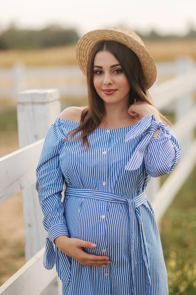 美丽的孕妇在蓝色的衣服和针织帽子站在附近的又懂法语在农场。她把手放在肚子上微笑着 — 图库照片
